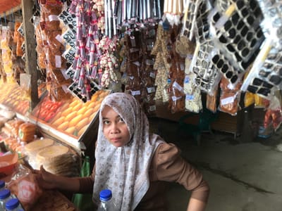 A woman wearing a white and beige patterned hijab and a brown long-sleeved top is the central focus, looking directly at the camera with a neutral expression. She appears to be seated or kneeling behind a market stall, with her right hand resting on a counter.

The setting is a vibrant market stall, heavily laden with various goods. In the foreground, on the counter near the woman, are several plastic bottles with blue caps and stacks of round, flat, light-colored crackers or flatbreads wrapped in clear plastic. Behind her, a display case exhibits rows of bright yellow and red fruits, possibly mangoes or similar produce.

The background is filled with an abundance of packaged food items, primarily snacks, hanging from overhead structures and shelves. These items are in clear plastic bags and include a variety of crispy, dried, or processed foods such as brown shredded or fried snacks, white and black crackers, and other confectionery. Text "BINTANG" is visible on some of these packages. To the far right, a green plastic chair and a broom are partially visible on the ground. The scene suggests a busy, daytime market environment in Panteepanah, Indonesia, where the woman is likely a vendor.