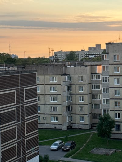 An aerial, eye-level view captures the evening sky over Kirishi, Russia, casting a warm, orange glow on apartment buildings and surrounding vegetation. The sky is partially obscured by streaks of pale clouds, suggesting the sun is near the horizon. In the foreground, a large, multi-story apartment building with dark, textured panels featuring a white geometric pattern dominates the left side of the frame. To its right, another, lighter-colored apartment building with a grid-like facade is visible, its windows appearing dark. Further in the background, a line of similar apartment buildings stretches across the landscape, interspersed with trees and topped with television antennas and radio towers. The ground level reveals a patch of green grass, a paved road, and two parked cars: a silver sedan and a dark SUV. A lone tree stands in the grassy area. The scene depicts a typical residential district at dusk.