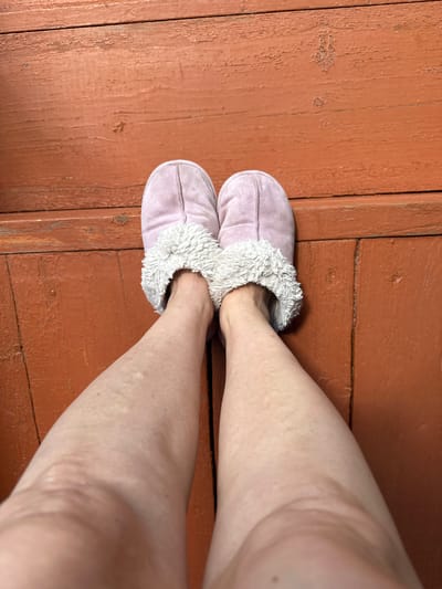 A pair of light pink slippers with white faux fur trim are worn by a person standing on a rustic, orange-painted wooden floor. The floorboards are worn and show signs of age and use, with visible grain and some peeling paint. The perspective is from above, looking down at the person's feet and lower legs. The person's legs are bare, and the skin appears fair. The scene is simple and domestic, suggesting a casual, indoor setting. The location is identified as Urazovo, Russia.