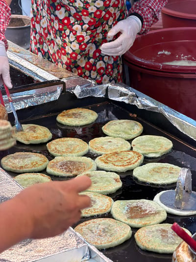 The image depicts a street food vendor in Jeju City, South Korea, preparing hotteok, a type of sweet Korean pancake, on a large, flat griddle. The griddle is covered in oil, and several hotteok are in various stages of cooking. Some are golden brown and slightly puffed, while others are still pale green and appear to be filled with a sweet mixture. The vendor, wearing a red and white checkered shirt and a floral apron, is using a pair of tongs to press down on one of the hotteok, indicating they are actively cooking. Disposable gloves are visible on the vendor's hands. In the background, large red plastic tubs, likely holding ingredients or finished products, are visible. The overall environment suggests a bustling outdoor market or street stall.