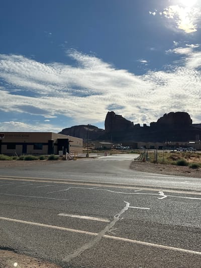 In Oljato-Monument Valley, United States, a wide asphalt road with white and yellow lines leads towards a low-lying building complex with a sign reading "UTAH FOOD BANK". In the background, a massive mesa, characteristic of Monument Valley, rises against a bright blue sky filled with scattered white clouds. The sun is visible in the upper right corner, casting a bright glare. The area appears to be a desert landscape with sparse vegetation. The overall scene is one of a functional, industrial building situated within a visually striking natural environment.