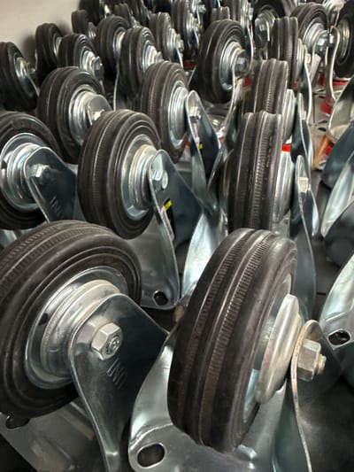 The image displays a close-up view of multiple industrial casters, also known as wheels. They are made of black rubber with silver metal mountings and hardware. The casters are arranged in rows and appear to be new or in very good condition. Some of the metal parts have labels or markings, such as "UNI" and what appears to be a yellow sticker. The background is out of focus and suggests an indoor environment, possibly a warehouse or storage area. There are no people visible in the image. The scene is static and depicts a collection of identical or very similar manufactured items. The location is not discernible from the visual information provided in the image.