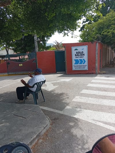 The image shows a street scene at what appears to be the exit of a school in Atamo Norte, Venezuela. A middle-aged man is sitting on a green plastic chair in the middle of the street. He is wearing a blue baseball cap, a white shirt, and dark pants. He is gesturing with his hands as if in conversation. Behind him is a red wall with a green door and a sign that reads "SÓLO SALIDA EXIT ONLY" with a security warning indicating that people are being filmed for safety. A crosswalk is visible in front of the wall. Trees are visible in the background, and a lamppost and camera are seen further back. The lower part of the picture shows the front of a motorcycle or scooter.