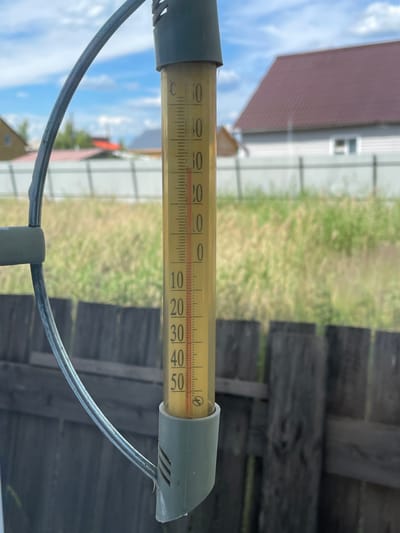 A vertical thermometer with a red liquid level is prominently displayed in the foreground, indicating a temperature of approximately 35 degrees Celsius. The thermometer is partially obscured by a curved metal rod, likely part of a weather station or an outdoor thermometer mount. In the background, out-of-focus elements suggest a rural or suburban setting. This includes a wooden fence, green vegetation, and the roofs of houses, one with a distinctive dark red, textured roof. The sky is partly cloudy, with visible white clouds against a blue backdrop. The scene appears to be captured in Yakutsk, Russia, a region known for its extreme temperatures, particularly its very cold winters. However, the temperature reading on the thermometer suggests a warm, possibly summer, day in Yakutsk.