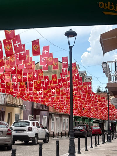 This image depicts a street scene in Cetinje, Montenegro, adorned with numerous small red flags bearing white crosses and crests. The flags are strung up, creating a festive or celebratory atmosphere, possibly for a national holiday or event. The street is lined with buildings, some of which appear to be historic, with balconies and awnings. There are streetlights and decorative black bollards along the sidewalk. Two cars are visible: a white Dacia with a Montenegrin license plate (PG CGP84) and a silver car parked behind it. The sky is visible with clouds, suggesting it is daytime, possibly a bright day given the lighting. The overall impression is a vibrant and decorated urban environment.