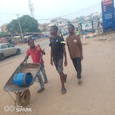 Three young males, likely boys or young teenagers, are walking along a light brown, possibly unpaved, road in what appears to be a busy town or village environment. The lighting suggests an overcast day or perhaps early morning/late afternoon, with soft, diffused light and no harsh shadows.
The boy on the left is pushing a dark-colored metal wheelbarrow that contains a blue metal drum or barrel lying on its side. He is wearing a red short-sleeved polo shirt, blue jeans, and dark footwear. His left hand is raised, possibly in a wave or gesture, and he has a neutral to slightly smiling expression.
Walking slightly ahead and to the right of the first boy is a second boy. He is wearing a dark (possibly black or very dark blue) short-sleeved t-shirt with a light-colored emblem or design on the chest, patterned shorts, and dark flip-flops. He is looking forward, slightly to his right.
The third boy is walking furthest to the right, slightly behind the second boy. He is wearing a brown short-sleeved polo shirt with a light design on the chest, dark trousers or long shorts, and dark slip-on footwear. He also has a neutral expression and is looking generally forward.
In the background, on the left side of the road, a light-colored car is driving past. Beyond that, there are makeshift stalls or market structures with tarpaulin roofs, indicating commercial activity. Further back, multi-story buildings and a tall metal communication tower are visible.
On the right side of the road, a large blue sign structure stands prominently. Text visible on this sign includes what appears to be a brand or business name, possibly "SARA" or "SAKI GARAGE" at the top, followed by "LUBRICANT," "AUTOCARE," and "MINI MART."
At the bottom left corner of the image, a "TECNO SPARK" watermark is present. The overall scene depicts everyday life and activity in a populated area.