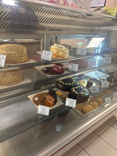 The image shows a display case filled with various cakes and pastries in a bakery. The bakery appears to be located in Samokov, Bulgaria, as suggested by a sign in Bulgarian that reads "При производствето на малотрайни сладкарски изделия се влагат продукти..." which translates to "For the production of perishable confectionery products, products are used...". This type of sign is common in Bulgarian food establishments.

Several cakes are visible, including a large, layered honey cake on the top shelf, a slice of layered cake with white cream and fruit on the top shelf, and a round red velvet cake. Below, there are chocolate cakes, some decorated with floral patterns and others with a more rustic appearance. Pastries, possibly croissants or similar baked goods, are also present on the bottom shelf. Price tags are visible for some of the items, indicating prices in Euros and Bulgarian Leva.

The display case is made of glass and metal, with shelves that appear to be wire racks. The lighting inside the case is bright, illuminating the baked goods. A person's reflection can be seen in the glass of the display case, suggesting that the photo was taken by someone standing in front of it. The background includes more shelving and what appears to be a calendar or notice board with dates. The flooring visible is tiled. The overall scene depicts a typical retail environment for a bakery.