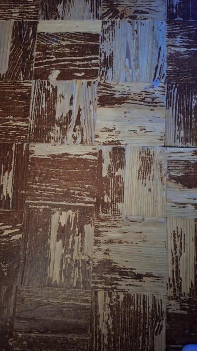 The image shows a close-up, top-down view of a wooden floor with a distressed, weathered, or intentionally aged finish. The flooring is composed of square tiles or planks arranged in a checkerboard pattern. Some tiles are a dark brown, while others are a lighter, bleached wood color, with the grain clearly visible. The surface of the wood exhibits a significant amount of wear and tear, with splintering, peeling, and gouges apparent. The texture appears rough and uneven. The lighting suggests an indoor environment, possibly artificial light casting some subtle blue tones in certain areas. The scene is devoid of people or any other discernible objects, focusing solely on the texture and pattern of the floor. There is no information to suggest this image is located in Lobnya, Russia; it is a generic depiction of a worn wooden floor.