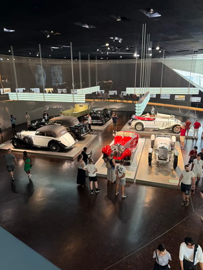 The image is a high-angle view of the Mercedes-Benz Museum in Stuttgart, Germany, showcasing a collection of classic cars. The museum features a modern architectural design with a dark ceiling, illuminated by track lighting and suspended light installations. The floor is polished dark wood, reflecting the lighting and the exhibits.
In the foreground, several people are observing the cars. Some are taking photographs with their mobile phones. A woman in a green dress walks across the floor, while a man in a grey t-shirt and shorts is partially visible on the left. Further into the scene, groups of visitors are gathered around various vintage automobiles.
The exhibits include several iconic Mercedes-Benz vehicles. A white convertible with its top down is prominently displayed, alongside a striking red convertible. To the left, a black convertible with a tan roof and a matching black sedan are parked. In the background, other vintage cars and a truck are visible, along with large projection screens displaying historical images and information. The overall atmosphere suggests a busy museum day with visitors engaging with the automotive history on display.