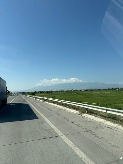 The image depicts a clear, blue sky over a highway. On the left, the side of a large, dark-colored truck is visible. The highway has multiple lanes, and other vehicles can be seen in the distance. To the right of the highway, a green field stretches out, bordered by a white metal guardrail with red reflectors. In the far distance, a large, snow-capped mountain range dominates the horizon, with a few clouds gathered around its peaks. The mountain is Mount Ararat, and the landscape suggests the image was taken near Arevabuyr, Armenia, given its proximity to the mountain. The lighting indicates it is daytime, likely late morning or early afternoon, with the sun casting shadows across the road. The scene is peaceful and expansive, showcasing a rural highway with a majestic natural backdrop.