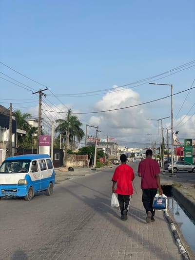 The image depicts a street scene in Ilado, Nigeria. Two individuals are walking along a paved sidewalk on the right. The first person is wearing a red shirt and black pants, carrying a blue bag. The second individual, closer to the camera, is wearing a red shirt and black track pants with white stripes, holding a white bag. A blue and white vehicle is parked on the street, facing the same direction as the pedestrians. Streetlights and power lines are visible overhead. Buildings and trees are situated along the street, including a Weima Bank signage. The sky is partly cloudy.