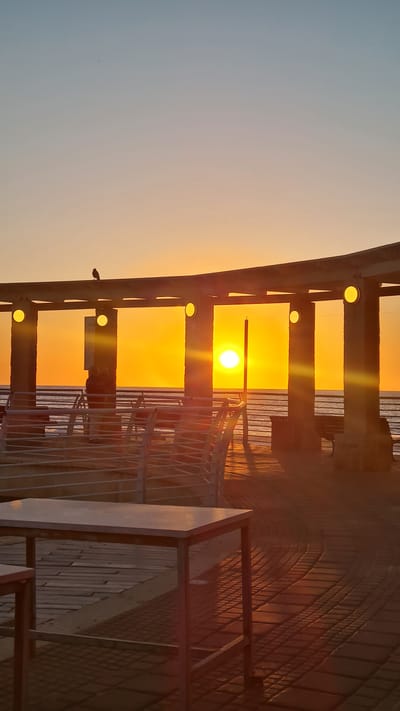 A horizontal view shows the setting sun. The sky on the left is pale blue that blends into the orange sky. The sun appears to be in the city of Nahariyya, Israel. A series of lights lead to the sun. A fence is on the right side of the image.