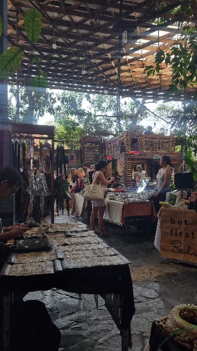 This image depicts a semi-outdoor market or craft fair located in Pecatu, Indonesia. The scene is illuminated by natural daylight filtering through a slatted wooden roof from which several bare light bulbs are suspended. Lush green foliage is visible around the perimeter and through openings in the roof structure. The ground consists of uneven, dark-toned paving stones.
In the foreground on the left, a person wearing glasses is partially visible, seated at a dark wooden table covered with numerous flat, possibly paper or textile, items. In the mid-ground, multiple market stalls are arrayed. To the left, racks display hanging items, appearing to be leather goods or textiles. Towards the center-right, a woman with dark hair, wearing light-colored shorts and a white top, stands facing a stall. She holds a mobile phone, directed towards the merchandise. She carries a white tote bag with visible text on its side.
Opposite her, behind the stall, stands a woman, likely a vendor, wearing a light-colored sleeveless top and glasses, observing the browser. The stall between them displays an array of small, reflective objects, consistent with jewelry, laid out on a light-colored cloth, with a small mirror also on the table. To the right of this stall, a cardboard sign reads "but first Bali".
In the background, an intricately carved, multi-tiered wooden structure is prominent, possibly serving as a large stall or decorative element. Other individuals are discernible in the background, moving through the market space.