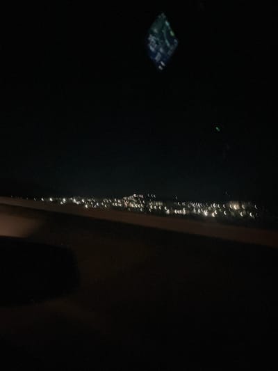 A night view shows a row of lights, likely from a building or structure, blurred and distorted, suggesting they were seen through a window with condensation or some other distortion. A small, blurry screen or display is visible on the left, reflecting light. The background is dark, and a portion of a wall or frame is visible on the right. The scene appears to be an interior view looking out at a lit structure at night, possibly in the city of Djerman, Bulgaria, although this is not definitively discernable from the image alone.