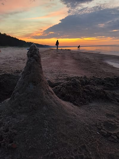 An outdoor, horizontal, medium shot captures a sandy beach scene during sunset. In the foreground, a large, elaborately constructed sandcastle dominates the frame, its textured surface detailed by the low-angle light. Beyond the sandcastle, the sandy beach stretches towards the ocean, with a dark, forested tree line visible in the distance on the left. Two silhouetted figures are present: one standing near the treeline on the left and another bending over near the water's edge on the right. The sky is a dramatic canvas of sunset colors, with hues of orange, yellow, and pink blending into soft gray clouds. The ocean is calm, reflecting the warm light of the setting sun. The overall impression is one of peaceful, late-day beach activity. This scene is located at the beach in Apšuciems, Latvia.