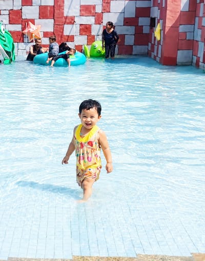 In Pantai Cermin Kiri, Indonesia, a young child stands in shallow, clear blue water at a water park. The child, wearing a yellow swimsuit with a colorful fruit pattern, smiles brightly and faces the camera. The water is clear, revealing a tiled bottom. In the background, several people are visible. A woman in a dark blue outfit stands near a green, cartoonish frog-shaped water feature. Further back, a group of people, including children, are in a blue inflatable ring. A red and white brick wall forms the backdrop of the water park, with a yellow star decoration attached to it. Green inflatable decorations are also present on the left side of the image. The scene depicts a sunny day at a family-friendly water attraction.
