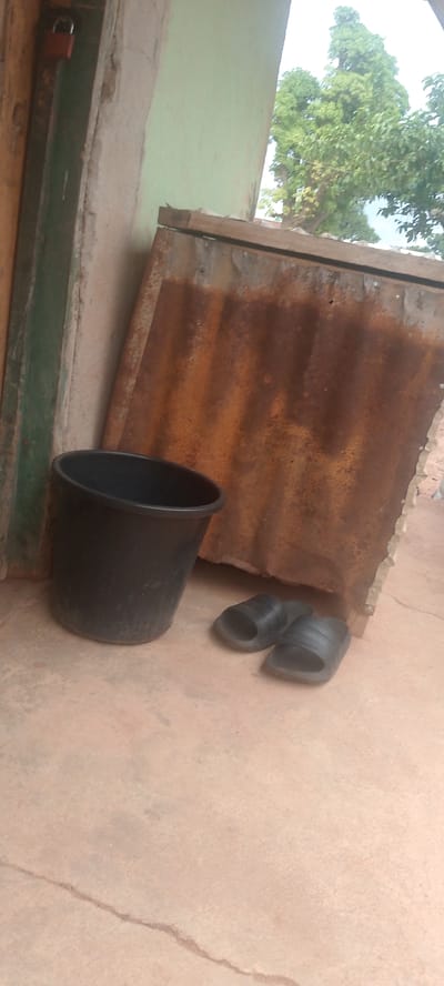 A black plastic bucket and a pair of black flip-flops sit on a dusty, reddish-brown concrete ground in front of a weathered, corrugated metal sheet. The metal sheet is rusty and leans against a rough, pale-green painted wall. To the left, a portion of a wooden door is visible, with a small, red padlock attached. The scene suggests an outdoor, possibly semi-enclosed area, common in a residential setting. The lighting indicates it is daytime, and the overall impression is one of simplicity and functionality. The concrete ground shows a crack running through it, hinting at a potentially dry or arid environment. In the background, lush green trees are visible against a bright sky, suggesting a tropical or subtropical climate. There is no discernible activity or event taking place; the objects appear to be casually placed. No text is visible in the image.