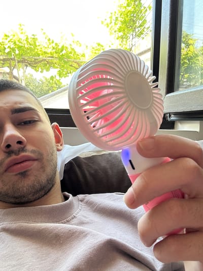 A young man is depicted in a close-up, rotated portrait, using a portable fan indoors. He has short dark hair, a beard, and a mustache, with a neutral expression. He is wearing a light-colored, possibly beige or grey, long-sleeved shirt. His right hand holds a white and pink portable fan, positioned in front of his face. The fan's pink blades are visible through its white cage, and a small blue indicator light illuminates near the base of the handle, suggesting it is active.
Behind the man, a large window reveals a vibrant outdoor scene with abundant green trees and foliage, indicative of bright daylight. The window has dark frames. A dark brown pillow or piece of furniture is partially visible behind his head. The setting suggests an indoor space, possibly a home, in Gotse Delchev, Bulgaria, where the individual is cooling himself with the fan on a bright day. There is no visible text in the image.