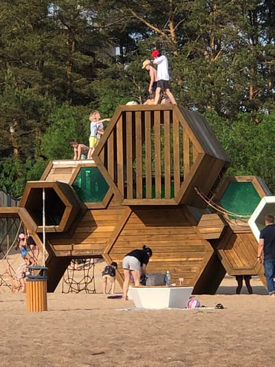 This image depicts an outdoor scene at the "Dune" eco-trail and children's playground in Sestroretsk, Russia, under bright daylight. The foreground and midground consist of sandy terrain, backed by a dense line of tall pine trees.
A large, multi-level wooden playground structure, constructed from interconnected hexagonal modules, dominates the center of the image. Several of these modules feature green transparent panels. Wooden railings secure the upper platforms of the structure, and rope climbing nets are visible on the lower left and beneath the central sections.
On the upper left platform of the playground, a young child with blonde hair, wearing a light-colored shirt and yellow shorts, is standing and pointing towards the right. Adjacent to them, a younger child, possibly a toddler, in a light-colored top, is on all fours, looking down. On the upper right platform, two male individuals are standing. One wears a red cap, a white long-sleeve shirt, and dark shorts, with a backpack partially visible. The other, wearing a light-colored cap and dark shorts (shirtless), stands nearby. Another person is partially visible lying down behind them.
At ground level on the left, a young child wearing a pink hat and a colorful swimsuit is on a rope net, looking to the right. A cylindrical, slatted wooden trash receptacle stands nearby. In the center-right foreground, a woman with dark hair, wearing a dark top and light-colored shorts, is leaning over a white rectangular basin or table. A plastic water bottle and a cup are placed on this basin. To her right, a man in a dark t-shirt and blue jeans stands with his back partially turned, facing right. On the sand near the white basin, a pink and white striped ball and dark sandals are visible. Partial views of human legs indicate individuals inside the lower hexagonal modules of the playground.
