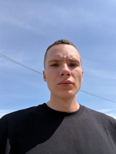 The image is a close-up, low-angle shot of a young man with short hair, looking directly at the camera. He is wearing a black t-shirt. The background consists of a clear blue sky with two parallel power lines stretching across it. The lighting suggests it is daytime, with sunlight casting a shadow on the man's t-shirt. The image provides no specific geographical markers or context to associate it with Amursk, Russia, or any other city.