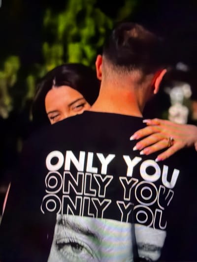The image shows a close-up of two people hugging, with the back of a black t-shirt prominently displayed. The t-shirt has bold white lettering that reads "ONLY YOU" repeated multiple times. The lower portion of the t-shirt features a black and white image of a woman's eye. A woman's hand with manicured nails and a ring is resting on the man's shoulder. The background is blurred and appears to be outdoors with greenery. The location in the city of Tomai, Moldova is not discernible from the image.