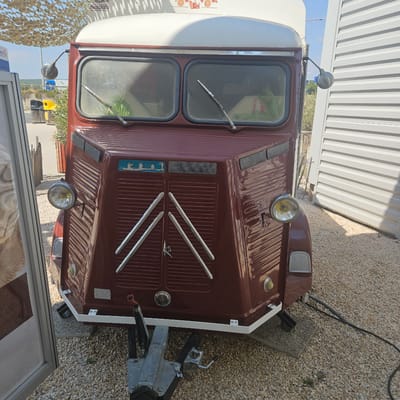 The image shows the front of a vintage, maroon-colored vehicle, possibly a food truck or camper, parked on a gravel surface. The vehicle has a distinctive ribbed front panel and two large windshields. Round headlights are mounted on either side of the front. A tow hitch is visible at the bottom. In the background, there is a white corrugated metal building, some vegetation, and a camouflage-patterned shade structure overhead. A blue and yellow trash bin is also visible in the mid-ground. The scene appears to be outdoors in a semi-industrial or commercial area. The image is taken from a frontal perspective, looking slightly upwards at the vehicle. There are no people visible in the image. The location is identified as the city of La Palme, France, suggesting it is a public space within that area.