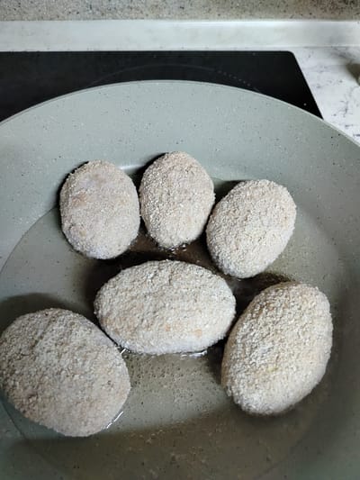 In a gray frying pan with oil, six breaded cutlets are being fried. The cutlets are oval-shaped and appear to be made of ground meat or fish, coated in breadcrumbs. The oil is shimmering and a few bubbles are visible, indicating the cutlets are actively cooking. The pan is set on a stovetop with a dark, possibly induction, cooktop visible behind it. The background shows a speckled countertop and a white backsplash. The overall lighting suggests an indoor setting, likely a kitchen, during the daytime. There is no visible text in the image. The scene depicts a food preparation activity, specifically the frying of cutlets.