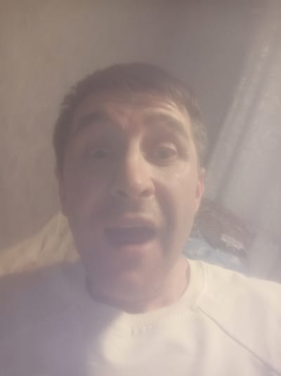 The image is a selfie of a man with short brown hair and light skin, wearing a white sweatshirt. He has wide eyes and his mouth is open in surprise. The background is blurry and appears to be indoors, with some fabric textures visible. The lighting is soft and diffused, creating a hazy effect over the image. The overall impression is of a candid, informal moment captured with a camera. There is no specific information in the image to determine the location within the city of Irbit, Russia.