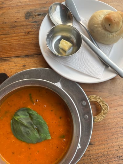 An overhead shot captures a rustic meal laid out on a wooden table. The primary subject is a vibrant orange-red soup, presented in a round, hammered metal pot. A large, fresh green basil leaf floats decoratively on the surface of the soup, which appears to contain flecks of other herbs and small oil bubbles. The metal pot features an ornate brass or bronze handle with intricate detailing.
To the left of the soup, a white ceramic plate holds a unique, light-brown bread roll. The bread is intricately shaped to resemble an animal, possibly a stylized turtle or a bird with a coiled body, and features a small dark dot that could be an eye. Alongside the bread, the plate holds two silver-colored metal utensils: a spoon and a small ladle. A tiny metal cup also sits on the plate, containing a small cube of light-yellow butter.
The meal is set on a warm-toned wooden table with visible grain and texture, suggesting a natural or rustic environment. The scene is brightly illuminated by natural daylight, indicating it is daytime. No people are visible in the image. The setting suggests a cozy meal experience, likely in a cafe or restaurant in Pokhara, Nepal.