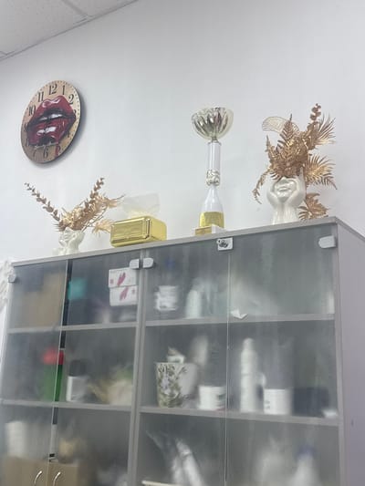 The image shows a close-up of a frosted glass-fronted cabinet in what appears to be a beauty salon or spa, likely located in Komsomolsk-on-Amur, Russia. The cabinet has two doors, each with two shelves, revealing various bottles, jars, and other products. Above the cabinet, on the wall, is a round clock with a lip design and the words "Beauty Studio." To the right of the clock, there are decorative golden foliage in a white vase and a golden tissue box. A trophy is also visible on top of the cabinet. The overall scene suggests a professional setting focused on beauty treatments.