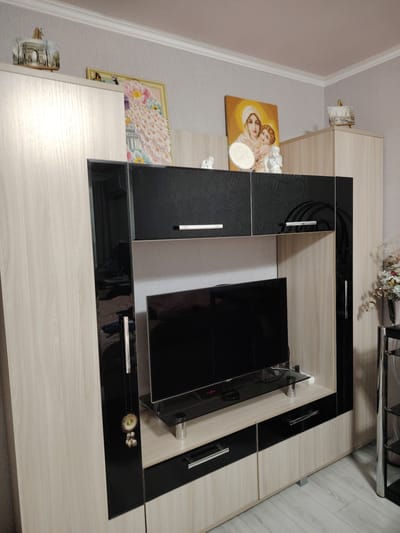 This is a medium shot of a living room wall unit in an apartment. The unit is made of light wood and black glossy material. It houses a black flat-screen television on a glass shelf. Above the television are black cabinets with embossed patterns and white handles. On top of the unit, there are several decorative items, including framed artwork depicting a peacock and a religious icon of Mary and Jesus, as well as small white angel figurines and a miniature structure resembling the Arc de Triomphe. A vase with artificial flowers is visible to the right. The wall behind the unit is a light textured wallpaper. The floor is light wood laminate. There are no people or animals visible in the image. Based on the context provided, this scene is located in an apartment in the city of Ussurijsk, Russia.