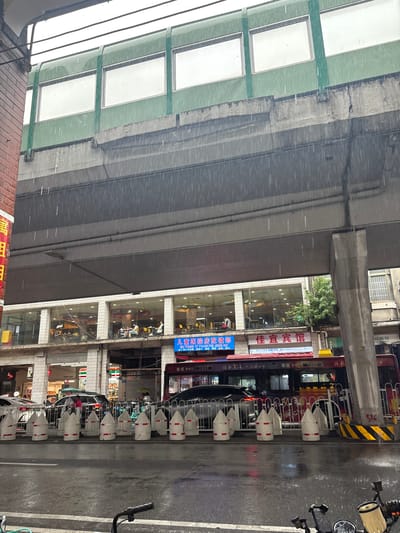 The image captures a street scene in Liwan District, China, during a heavy rainfall. A multi-level structure, likely an elevated walkway or road, dominates the upper portion of the frame, with concrete supports extending downwards. Beneath this structure, a row of businesses with large glass windows is visible, showcasing various interiors and patrons. People are seated inside, some appearing to be dining or socializing.

At street level, the road is wet and reflects the surrounding lights. A line of white traffic barriers with red markings is positioned along the side of the road. Several vehicles are present, including cars and a bus. The bus is red and features advertising banners. A motorcycle is partially visible in the foreground, suggesting it is parked or being operated by the photographer. The rain is clearly visible as streaks falling from above, creating a sense of a wet and overcast day. The environment appears to be urban, with the presence of buildings, signage, and transportation infrastructure.