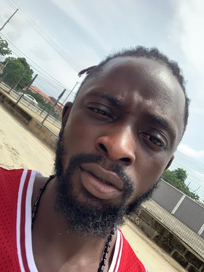 A close-up, angled selfie-style portrait features a Black man outdoors. He occupies the majority of the frame, looking directly towards the camera with a neutral to slightly pensive expression. His skin, particularly on his forehead, nose, and cheekbones, glistens, suggesting warmth or humidity. He has short, dark, tightly curled hair and a full, dark beard. He wears a red sleeveless jersey with white striped trim on the collar, and a dark, beaded necklace around his neck.
The background is an outdoor setting, likely under an overcast but bright sky during the daytime. Behind him, a chain-link fence is visible, partially obscuring what appears to be a light-colored building wall and some distant green trees. A glimpse of a white car can be seen beyond the fence. The ground in the foreground and mid-ground appears to be dirt or sand. The overall scene suggests an urban or semi-urban environment in Sangotedo, Nigeria. There is no visible text.