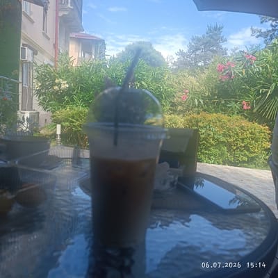 The media file depicts a close-up of a plastic cup filled with a beverage, likely an iced coffee, resting on a glass-top table. The cup is topped with a clear plastic dome lid and a black straw. In the background, a lush green garden with pink flowers and trees is visible under a bright blue sky with scattered clouds. A building with beige walls and a red-tiled roof is partially visible on the left. The scene suggests a casual outdoor setting, possibly a cafe or a patio. The time stamp at the bottom right indicates "06.07.2026 15:14", suggesting it was taken on July 6, 2026, in the afternoon. The lighting indicates it is a sunny day. The location is identified as Dagomys, Russia.