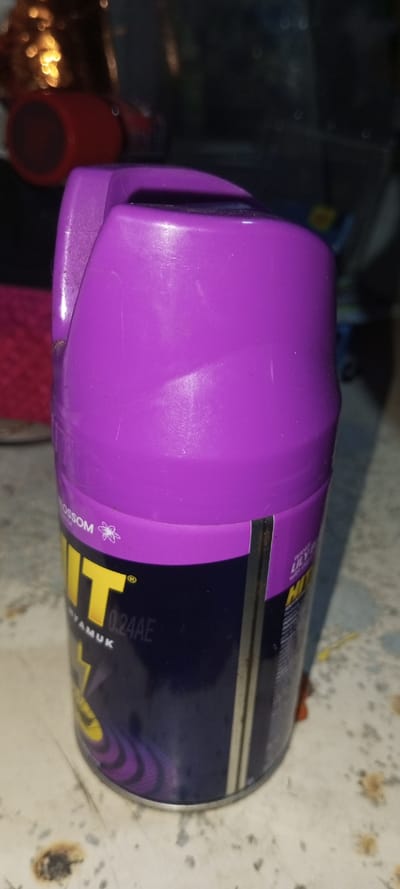 A purple Hit mosquito spray can stands upright on a speckled surface. The can has a purple top with a handle and a blue and yellow label. "Hit" is written in large yellow letters. The label also features purple swirling designs. The background includes a red object and other blurry, indistinct items. The surface the can is on appears to have small stains or spots.