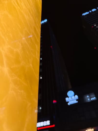 The image, captured at night in Huangpu District, China, features a close-up of a textured, yellow surface on the left, with a blurred cityscape in the background. The yellow surface appears to be illuminated from within or behind, revealing a marble-like pattern. To the right, tall buildings rise into the darkness, their lights creating streaks of color. Notably, there is a blue, stylized logo or sign visible on one of the buildings, and a red neon sign with Chinese characters also appears illuminated at the base of a structure. The overall impression is an urban night scene, with a focus on artificial lighting and architectural elements. There are no discernible people or specific actions taking place.