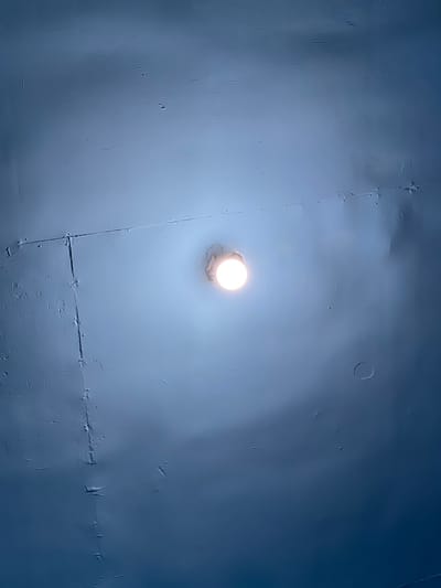 The image is an eye-level, medium shot of a blue-painted ceiling with a single, lit bulb centrally located and slightly to the left. The ceiling appears to be constructed from large panels, indicated by faint seams and rivets across its surface. The paint has a matte finish and exhibits subtle imperfections, such as minor cracks, bumps, and what appear to be small water stains or patches of uneven texture. The light from the bulb creates a soft, diffused glow, illuminating the immediate area and casting gentle shadows that highlight the ceiling's textures. The overall impression is one of a utilitarian interior space. There is no visible information in the image to suggest the location in the city of Bandarbireuen, Indonesia.