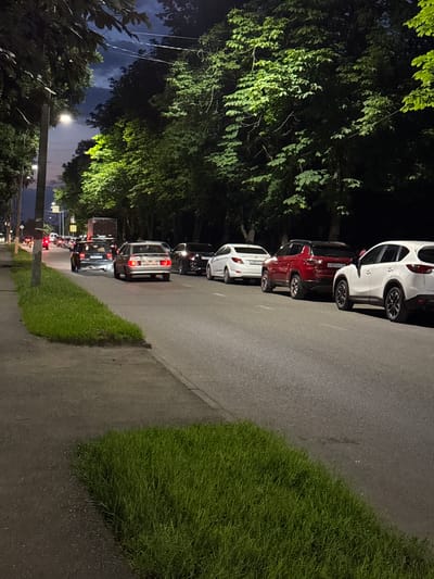 The image depicts a street scene at night in Vladikavkaz, Russia, with cars parked along the side of a road. The lighting suggests it is nighttime, with artificial streetlights and car headlights illuminating the scene. The road is paved, and a strip of lush green grass borders it on the right side of the frame. Tall trees with dense foliage are visible on the left, their leaves illuminated by a light source, casting shadows.

Several cars are parked in a line, one after another, on the side of the road. The cars appear to be a mix of makes and models, with visible license plates on some. The car closest to the foreground is white, followed by a red car, and then several other vehicles further down the road. Brake lights are visible on some of the cars, indicating they are either parked or stopped in traffic.

The overall atmosphere is calm and quiet, typical of a residential street at night. There are no people visible in the immediate vicinity of the cars, suggesting they are parked for the night or their occupants are inside nearby buildings. The sky is dark, with hints of clouds and a distant light source, possibly street lighting or ambient city glow. No specific event or activity is clearly taking place, beyond the passive presence of parked vehicles on a street. The scene is set in a suburban or urban environment, characterized by the presence of roads, trees, and parked cars.