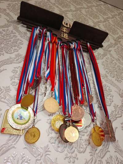 This is a medium-shot, eye-level photograph of a wooden medal rack mounted on a patterned wallpapered wall. The rack is dark brown and has several slots for holding ribbons. Attached to the rack are numerous medals, most of which have red, white, and blue ribbons, suggesting they are from Russian competitions. Some medals are gold, silver, or bronze colored, and several have visible inscriptions or images. One medal in the lower left displays an image of two individuals engaged in a judo match, with text that appears to be in Russian and reads "МЕСТО" (Mesto), meaning "place" or "ranking". Another medal displays a prominent number "3" with stars around it, likely indicating a third-place finish. The background wallpaper has a textured, floral-like pattern in a light, neutral color. The photograph does not depict any people or specific geographical landmarks that would indicate its location within the city of Zlatoust, Russia.