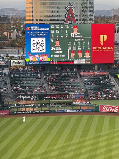 The image captures a baseball game in progress at Angel Stadium in Anaheim, California, United States. The focus is on the stadium's large video screen, displaying team statistics and advertisements. On the left side of the screen, a blue advertisement with the text "ANGELS. OCNAVIGATOR.ORG" promotes mental wellness resources and includes a QR code. To the right, baseball statistics for players like Randal Grichuk and Sam Alldegheri are shown. A red advertisement for Pechanga Resort Casino occupies the right side of the screen.

Below the video screen, spectators are seated in the stands, watching the game. Advertisements from companies such as Modelo, Nitto Tire, and 714Tickets line the stadium walls. On the baseball field, a single player in a white uniform stands on the field. The green grass is precisely striped. Palm trees and buildings can be seen in the background, suggesting the stadium's urban setting.