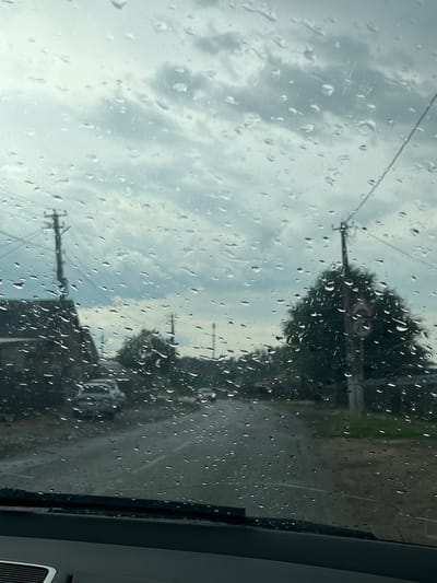 The image depicts a view through a rain-streaked windshield of a car parked on a paved road in Leningradskaya, Russia. The sky is overcast with heavy, grey clouds. On the left side of the road, a house with a dark roof and windows is visible, with utility poles and wires in front of it. Further down the road, a white car is parked. On the right side, a large tree stands next to a utility pole with a circular road sign depicting a number 70 inside a red border, indicating a speed limit. The road appears wet and possibly muddy, suggesting recent or ongoing rainfall. The overall impression is a somber, rainy day in a semi-rural or suburban setting.