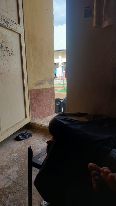 The image depicts an interior scene looking out through an open doorway. The interior features a worn, pale yellow wall with some peeling paint and a textured reddish-brown lower section. To the left, a light-colored, distressed metal door is ajar. In the foreground on the right, a person is seated, with their lower body and feet visible, covered by dark fabric. A dark plastic bag lies on the concrete floor to the left, near the doorway.
Looking through the doorway, a building with a light green roof is visible, suggesting an outdoor area in front of it. Several black buckets are lined up on the ground outside. Clothes are hanging on a line in the background, and a person in a blue shirt can be seen near the building. The ground outside appears to be concrete or paved. The overall environment suggests a residential or institutional setting in New Nyanya, Nigeria.