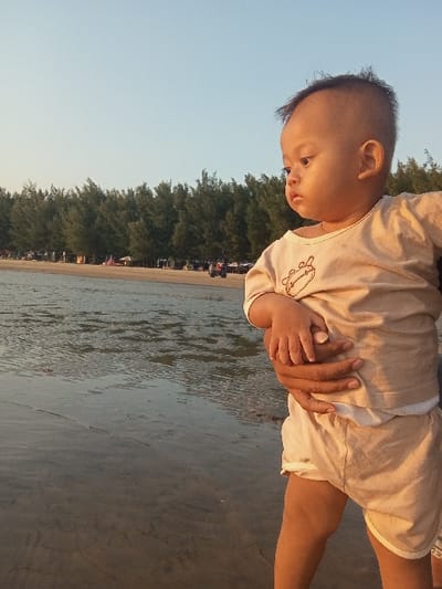 An outdoor, eye-level photograph depicts an infant held by an adult on a beach in Tritunggal, Indonesia, under a clear, light blue sky suggesting late afternoon or early evening. The infant, with light-toned skin and a short, dark haircut, wears a light beige long-sleeved shirt adorned with a dark animal graphic and matching shorts. The child's head is turned towards the left, observing the distant scene. A dark-skinned adult's right hand and forearm are visible, supporting the infant's lower torso. The foreground shows shallow, rippled water receding from a wet sandy shore. In the mid-ground, a dense horizontal line of tall, slender evergreen trees separates the beach from a further background area. Beyond the tree line, indistinct human figures, small structures, and a single vehicle are faintly discernible on the sandy terrain.