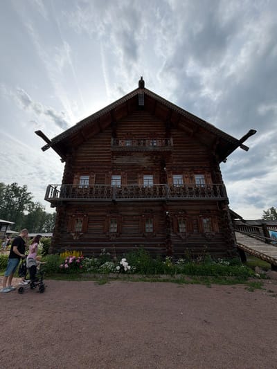 In Dnp Ryzhiki, Russia, a large, ornate wooden house with a steep roof and intricate carvings stands under a cloudy sky. The house has two balconies and multiple windows with wooden shutters. In the foreground, a man in a black t-shirt and denim shorts stands with his arm around a woman in a pink shirt. A child is in a stroller next to them, with the child reaching out towards a flowerbed. The flowerbed contains pink and yellow flowers and greenery. The ground in front of the house is covered in gravel. A wooden ramp leads up to the right side of the house.