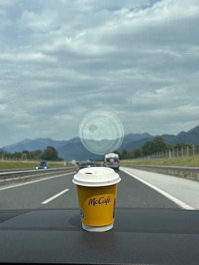 A McCafe coffee cup is placed on the dashboard of a vehicle, viewed from inside the car. The cup is yellow with red and white branding. Through the windshield, a highway is visible with cars in motion. In the distance, rolling hills and mountains are seen under a cloudy sky. The view through the windshield suggests the vehicle is traveling on a highway in Hraše, Slovenia, based on the presence of a typical European highway layout and the distant mountain ranges resembling the Slovenian landscape. There is a circular reflection on the windshield, likely from a sun visor or other interior object.