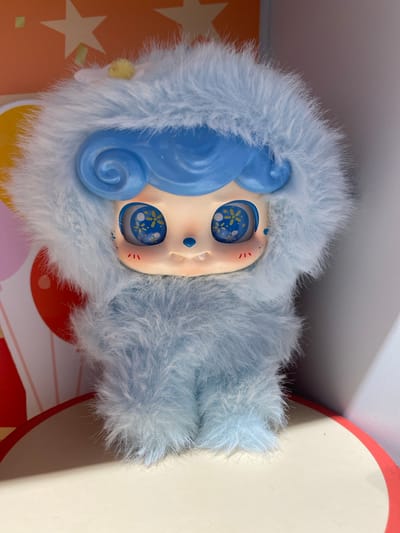 A light blue, furry doll with a blue swirl hairdo is standing on a round surface with a red border. The doll has large, round eyes with blue irises containing yellow floral patterns and blue specks. Its cheeks have faint red marks, and its mouth is open slightly, revealing small white teeth. A small blue nose is positioned centrally on its face. The doll is covered in light blue fur, giving it a fluffy appearance. In the background, there are colorful shapes, including a yellow star and pink and red circles, on an orange and white patterned surface. The scene is lit by artificial light, casting soft shadows. This image was taken in East Jakarta, Indonesia.