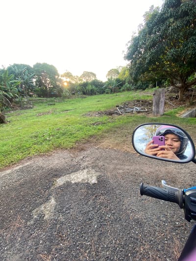 The image captures a reflection in a motorcycle's rearview mirror, showing a young person taking a selfie. The person is wearing a helmet and a dark hooded jacket, holding a purple smartphone up to take the photo. They appear to be smiling. The scene behind them, visible in the mirror and the wider image, is an outdoor, rural setting. Lush green grass covers a sloping field, with various trees and banana plants on the left. A wooden post and some scattered debris are also visible in the background. The sun appears to be low in the sky, casting a warm glow and creating lens flare through the trees, suggesting it is either early morning or late afternoon. The ground in the foreground is a rough asphalt surface with small stones and dry twigs, indicative of a road or pathway. The overall mood is casual and relaxed, with a hint of the tropical environment suggested by the vegetation and the time of day. There is no visible text in the image. The location context provided is Banjarbaru, Indonesia.