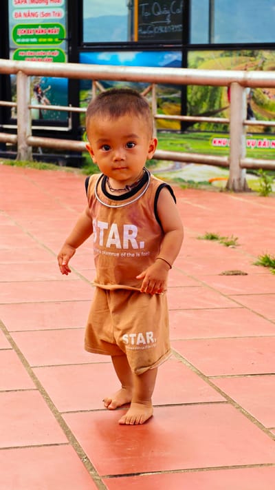 A toddler of East Asian ethnicity stands barefoot on a reddish-orange tiled ground, looking directly at the camera. The child wears a light brown sleeveless top and matching shorts. The top is emblazoned with "STAR." in white capital letters, with smaller, lighter text underneath reading "Whteie plamrue of the is font". A black string necklace with a brown charm and a silver-colored rigid collar encircles the child's neck, and a thin brown string bracelet adorns the right wrist. Behind the child, a light gray metal railing extends horizontally across the midground. In the background, a building with large glass windows reflects or displays posters of lush green terraced landscapes and mountains, indicative of the region around Tu Le, Vietnam. To the far left, a green sign with white Vietnamese text reads "SAPA MƯỜNG HUA" and "ĐÀ NẴNG (Sơn Trà)", followed by "ĐIỂM DỪNG CHÂN NGHỈ NGƠI". Above the right side of the railing, a dark sign with white Vietnamese text partially visible as "Tụ Quây Cổ" and "Hạn 098" is affixed to the building. Sparse green vegetation is visible beyond the railing.