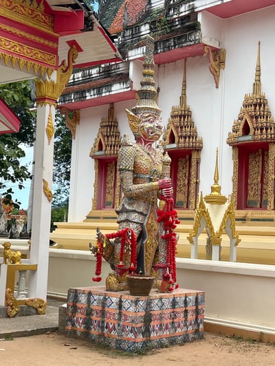 This is a medium shot, eye-level, outdoor photograph of a statue in front of a white building with red and gold trim. The statue is of a large, humanoid figure with a red face, intricate gold armor, and a tall, ornate headdress. Red garlands are draped around the statue's neck and arms. The statue stands on a decorative pedestal with a patterned base. Behind the statue, the building features several windows with gold trim and red interiors. The roof of the building is tiled and appears weathered, with some greenery growing from it. The ground in front of the statue is sandy and there are some fallen leaves. To the left of the statue, a white pillar with gold decorative elements is visible. The overall scene suggests a religious or cultural site. The location is identified as Baan Harn, Thailand.