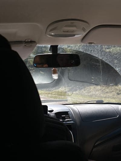 The image captures a bright daytime scene from inside a vehicle, looking forward through the windshield. On the left, parts of the vehicle's interior are visible, including a light-colored overhead console with integrated lights or buttons, and a sun visor. A dark dashboard with air vents and other controls fills the lower-right portion of the frame, suggesting a sturdy vehicle interior, possibly a van or truck.
The central focus is the rearview mirror, which reflects a person's face, likely the driver's, and a smartphone, possibly mounted on the dashboard. A small white object, resembling a charging adapter or sensor, is attached to the windshield near the rearview mirror. The windshield itself appears somewhat dusty or dirty.
Beyond the windshield, a sunny outdoor environment is visible. The ground immediately outside the vehicle shows a dirt or gravel road with some sparse vegetation. In the distance, the front of another vehicle, possibly a van, is partially discernible. The clear, bright light indicates a sunny day. No specific activity is evident beyond being inside a stationary vehicle. There is no visible text. The overall setting suggests a rural or semi-rural location, consistent with Gol'yany, Russia.